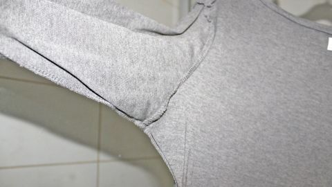 Du kannst Schweißflecken und Schweißgeruch vermeiden, indem du eine Tagesslipeinlage in der Mitte durchschneidest und in das Achselteil deines Kleides klebst. Ein graues Shirt hängt an einem Kleiderbügel, ohne sichtbare Schweißflecken oder Geruch, in einem hellen Raum.