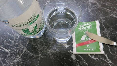 Ein Glas mit Mineralwasser steht neben einer Flasche mit der Aufschrift "Natürliches Mineralwasser medium" und einem Päckchen "Kaiser-Natron" auf einer Marmoroberfläche. Ein Messer liegt auf dem Päckchen.