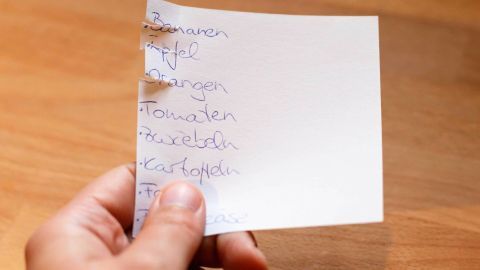 Eine Hand hält einen Zettel mit einer Liste von benötigten und bereits vorhandenen Dingen in einer Küche.