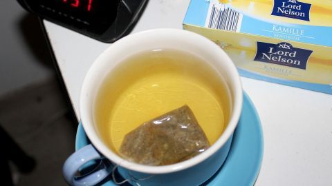 Ein Schälchen mit aufgegossenen Kamillenblüten im Schlafzimmer lindert nächtliche Hustenanfälle. Eine Tasse mit Kamillentee steht auf einem Nachttisch. Der Teebeutel zieht in der warmen Flüssigkeit, während die Uhr 21:51 anzeigt.