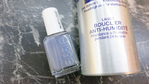 Ein Fläschchen mit lila Nagellack steht neben einer Dose Haarspray auf einer marmorartigen Oberfläche.