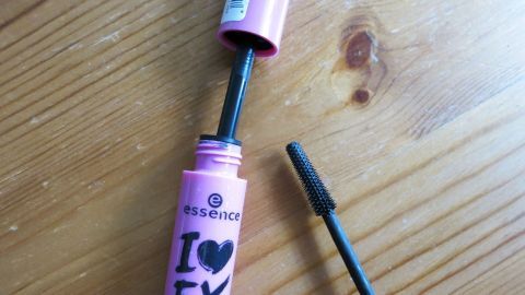 Mithilfe eines alten Wimperntuschebürstchens ein Verklumpen der Wimpern beim Tuschen der Wimpern verhindern. Eine geöffnete Wimperntusche liegt auf einem Holzuntergrund, während die Bürste bereit ist, verklumpte Wimpern zu bösen.