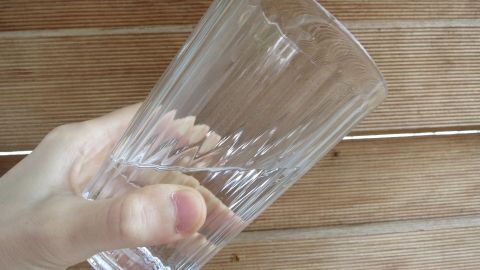 Mit dieser Methode besiegt man Schluckauf: Ein Glas Wasser am gegenüberliegenden Rand mit den Lippen berühren und damit quasi umgekehrt vornübergebeugt langsam ein paar Schlucke trinken. Danach wieder aufrichten. Ein Hand hält ein Glas Wasser, während es verkehrt herum geneigt wird, auf einer Holzoberfläche.