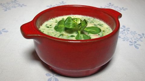 Eine rote Schüssel enthält Feldsalatsuppe, garniert mit frischem Feldsalat, auf einem Tisch mit dekorativem Muster.