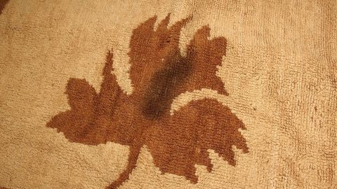 Ein brauner Fettfleck auf einem hellen Teppich zeigt eine unregelmäßige Form, die ein Blattmotiv ähnelt.