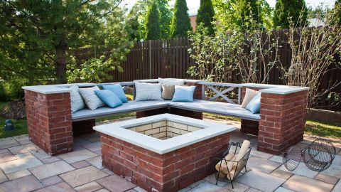 Eine Terrasse aus Betonsteinen mit einer Loungebereich und einem zentralen Feuerplatz, umgeben von Bäumen und Hecken.