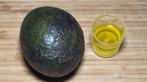 Glänzende und kräftige Haare bekommt man, wenn man sie mit Avocado und Olivenöl pflegt. Eine Avocado liegt neben einem kleinen Glas mit Olivenöl auf einer Holzoberfläche, bereit zur Haarpflegeanwendung.