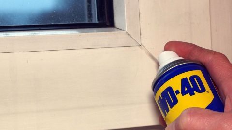 Eine Hand hält eine WD-40-Spraydose und entfernt Klebereste von einem Fensterrahmen in einem hellen Raum.