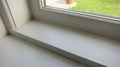 Mit Nagellackentferner kann man Rußflecken vom Fensterrahmen erfolgreich entfernen. Vorher an einer nicht so gut sichtbaren Stelle ausprobieren, ob der Kunststoff die Behandlung unbeschadet übersteht. Ein sauberer Fensterrahmen wird gereinigt, um Rußflecken zu entfernen, während ein blühender Garten im Hintergrund sichtbar ist.