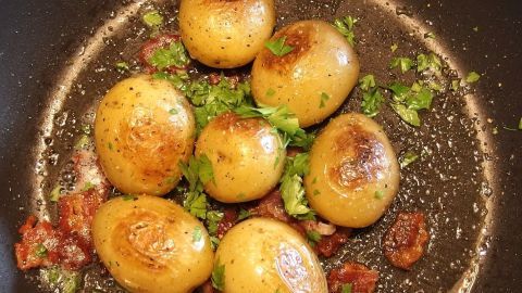 Kleine Frühkartoffeln bräunen in einer Pfanne mit frischer Petersilie und Speckstücken, umgeben von einer goldbraunen Kruste.