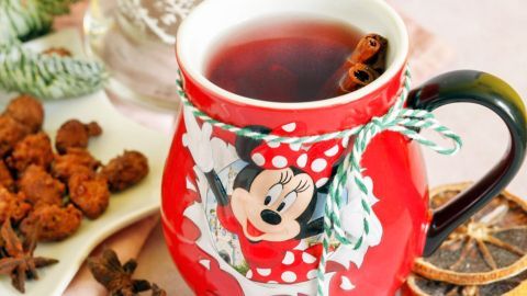 Ein roter Kinderpunsch in einer Minni-Maus-Tasse dampft neben Gewürzen und Trockenfrüchten auf einem Tisch.