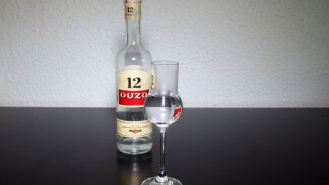 Eine Flasche mit klarer Flüssigkeit steht neben einem Glas, das zur Hälfte mit dem Inhalt gefüllt ist, auf einem dunklen Tisch.