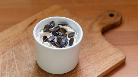 Ein kleiner, weißer Becher mit Kürbiskernaufstrich steht auf einem Holzbrett, dekoriert mit Kürbiskernen.