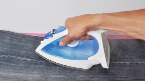 Mit dem DIY-Trick zum Bügeln: Mach dir das Bügeln leichter! Verwende eine Mischung aus Wasser und Essig, um hartnäckige Falten in deiner Lieblingsjeans mühelos zu glätten. Ideal für effiziente Bügelarbeit. Eine Hand bügelt mit einem blauen Dampfbügeleisen eine Jeans auf einem rosa Bügelbrett im Kontext von Hausarbeit.