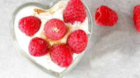Ein herzförmiges Glas enthält cremigen Mascarpone-Himbeerquark, garniert mit frischen Himbeeren und einer Prise Keksbrösel.