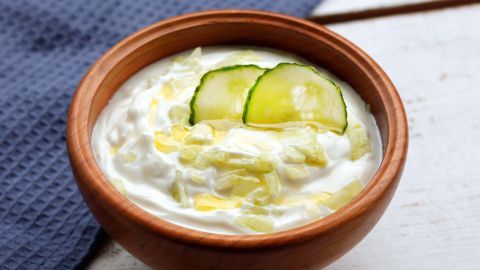 Für dieses leckere Tzatziki-Rezept benötigt man Quark, eine Salatgurke und Knoblauch. Das Ganze wird anschließend mit Salz verfeinert. Eine Schale Tzatziki mit Stückchen von Gurke und einem Hauch Olivenöl steht auf einem Holzgrund, umgeben von einem blauen Tuch.