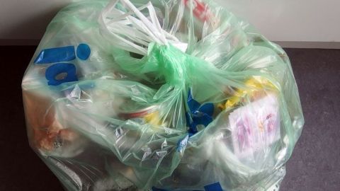Ein grüner Abfallsack, gefüllt mit verschiedenen Abfällen, steht auf einem dunklen Untergrund in einem Raum.