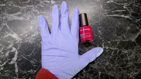 Mithilfe eines Einweghandschuhs lassen sich schlecht lackierte Fingernägel einfacher "reparieren". Eine Hand mit blauen Handschuhen liegt auf einem Tisch, während daneben eine Flasche Nagellack in leuchtendem Pink steht.
