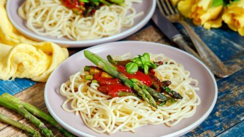 Pasta mit grünem Spargel und Tomatensauce liegt auf einem Teller, umgeben von frischem Gemüse und einem rustikalen Tisch.