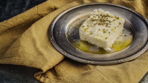 Ein Stück gegrillter Feta liegt auf einem silbernen Teller, umgeben von Olivenöl, auf einem goldenen Tuch.