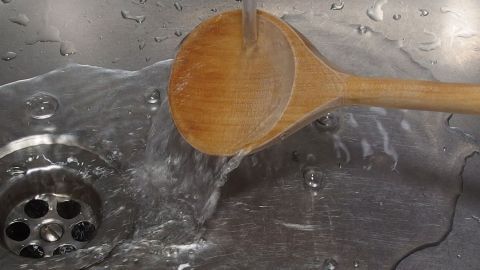 Ein Holzlöffel wird unter fließendem Wasser gewaschen, während er in einem Spülbecken mit Abflusslöchern liegt.