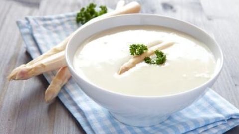 Eine Schüssel Spargelcremesuppe steht auf einem blau karierten Tuch, garniert mit Petersilie, daneben liegen frische Spargelstangen.