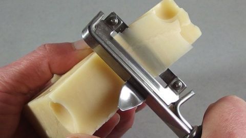 Ein Käse wird mit einem Sparschäler in dünne Scheiben geschnitten, gehalten von einer Hand auf grauem Hintergrund.