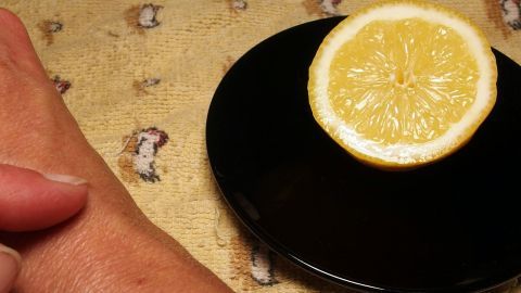 Eine Hand mit Altersflecken liegt neben einer halben Zitrone auf einem schwarzen Teller, umgeben von einem hellen, gemusterten Untergrund.