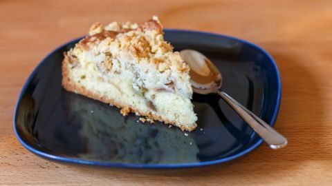 Ein Stück Rhabarberkuchen mit Streuseln liegt auf einem blauen Teller, während ein kleiner Löffel daneben ruht.