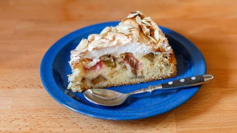 Ein Stück Rhabarberkuchen mit Baiser-Mandelhaube liegt auf einem blauen Teller neben einem Löffel auf einer Holzoberfläche.