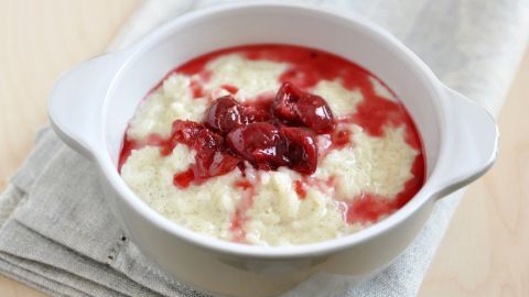 Eine Schüssel Milchreis mit einer fruchtigen Kirschsoße steht auf einem Tisch, umgeben von einem hellen Tuch.