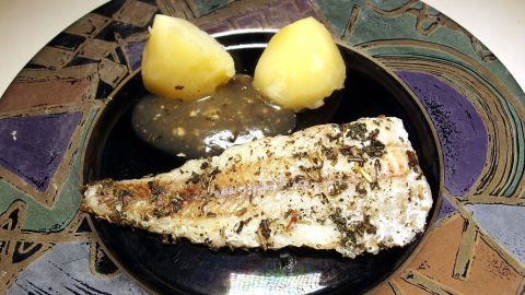 Gegrillter Fisch liegt auf einem schwarzen Teller, umgeben von zwei Kartoffeln und einer Sauce, serviert auf buntem Geschirr.
