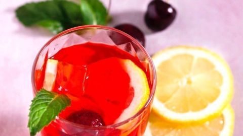 Ein Glas Sangria mit roten Früchten, Zitrone und Minze steht auf einem lichtrosa Hintergrund, umgeben von Kirschen und Zitronenscheiben.