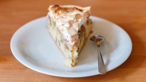 Eine Stück Rhabarberkuchen mit einer goldbraunen Baiserhaube liegt auf einem weißen Teller neben einem kleinen Löffel.