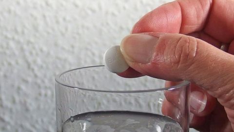Warmes Wasser zum Schlucken von Tabletten bei Halsschmerzen verwenden. Eine Hand hält eine Tablette über ein Glas Wasser, bereit zur Einnahme gegen Halsschmerzen mit schmerzhaften Schluckbeschwerden.