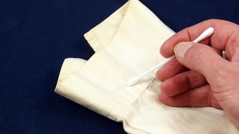 Eine Hand benutzt ein Wattestäbchen, um Flecken auf einem weißen Textil auf einem dunklen Hintergrund zu behandeln.