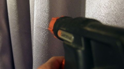 Ein Handgerät sprüht auf einen Vorhang, um Falten zu entfernen, in einem hellen Raum mit grauen Vorhängen.