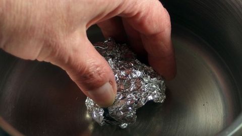 Als Ersatz für einen Topfreiniger, kann man zu Scheuern von Töpfen, auch Alufolie verwenden. Eine Hand greift in einen Topf mit Wasser, während sie ein Stück Alufolie zum Scheuern verwendet.