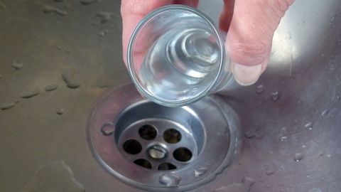 Ein Glas wird über einem Abfluss gehalten, wobei Wasser hineinfließt, um mögliche Verstopfungen zu lösen.