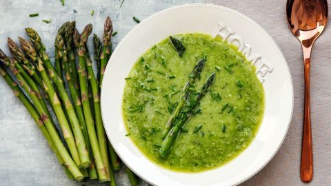 Eine grüne Spargelsuppe liegt in einer weißen Schüssel, umgeben von frischem Spargel und einem goldenen Löffel.