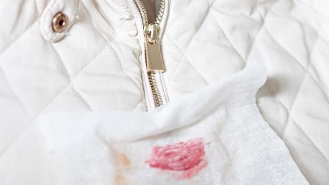 Ein Reinigungstuch entfernt Make-up-Flecken von einer weißen Jacke auf einem Holztisch.