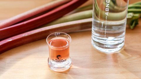 Ein Glas mit Rhabarberlikör steht auf einem Holztisch, neben einer Flasche und frischen Rhabarberstangen.