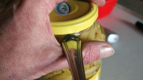 Eine Hand hält einen Glasdeckel mit einem Löffel, um ihn leicht zu öffnen, während er auf einer Tischoberfläche liegt.