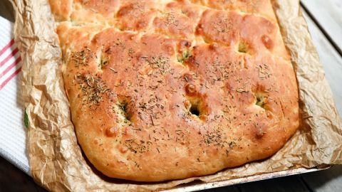 Ein frisch gebackenes italienisches Focaccia-Brot liegt auf gebräuntem Backpapier und ist mit Kräutern bestreut.