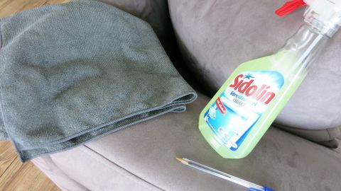 Ein graues Mikrofasertuch liegt auf einem Sofa, daneben steht eine Reinigungslösung und ein Kugelschreiber.