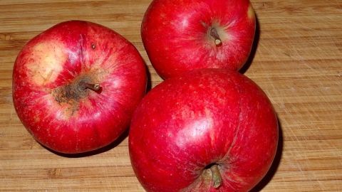 Mithilfe von Apfelsaft können geschälte Äpfel vom braun werden verschont werden. Drei rote Äpfel liegen auf einem Holzbrett. Die Äpfel wirken frisch und unverletzt, ideal für Apfelsaft.