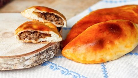 Goldbraune Empanadas liegen auf einem Tuch, eine halbierte zeigt die herzhafte Füllung aus Fleisch im Hintergrund.