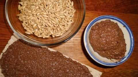 Eine cremige Schoko-Hafer-Creme liegt auf einer Brotoberfläche, daneben sind Haferflocken und eine kleine Schüssel mit Creme.