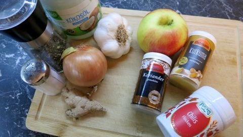 Auf einem Holzbrett liegen Zutaten für Apfel-Zwiebel-Schmalz (vegan), darunter eine Zwiebel, ein Apfel, Gewürze und Knoblauch.