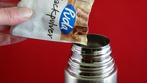 Eine Hand hält ein Päckchen Backpulver über eine Thermoskanne, bereit, es hineinzuschütten, vor rotem Hintergrund.